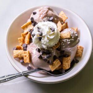 A bowl of ice cream topped with cereal squares, chocolate chips, whipped cream, and white sprinkles, with a spoon in the bowl.