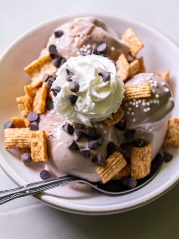 A bowl of ice cream topped with cereal squares, chocolate chips, whipped cream, and white sprinkles, with a spoon in the bowl.