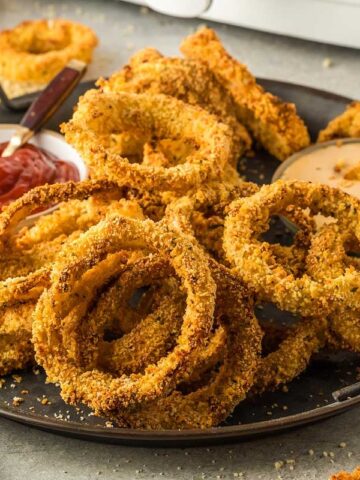 A plate of crispy, golden onion rings served with two dipping sauces, one ketchup and one mayonnaise-based, placed on a gray surface—just one of 15 air fryer side dishes that'll elevate any meal.