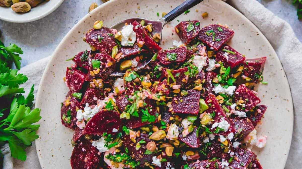 A plate of beet salad with crumbled feta cheese, chopped nuts, and fresh herbs. A serving spoon rests invitingly in the fresh salad. Fresh parsley is seen next to the plate.