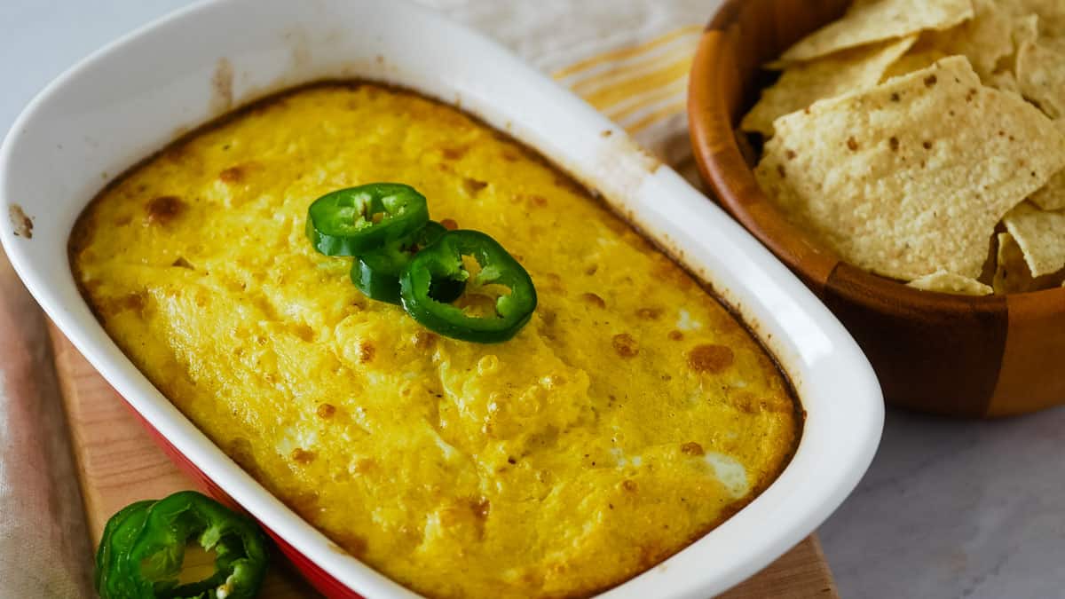 A baked jalapeño cheese dip in a white casserole dish, garnished with sliced jalapeños, next to a wooden bowl filled with tortilla chips.