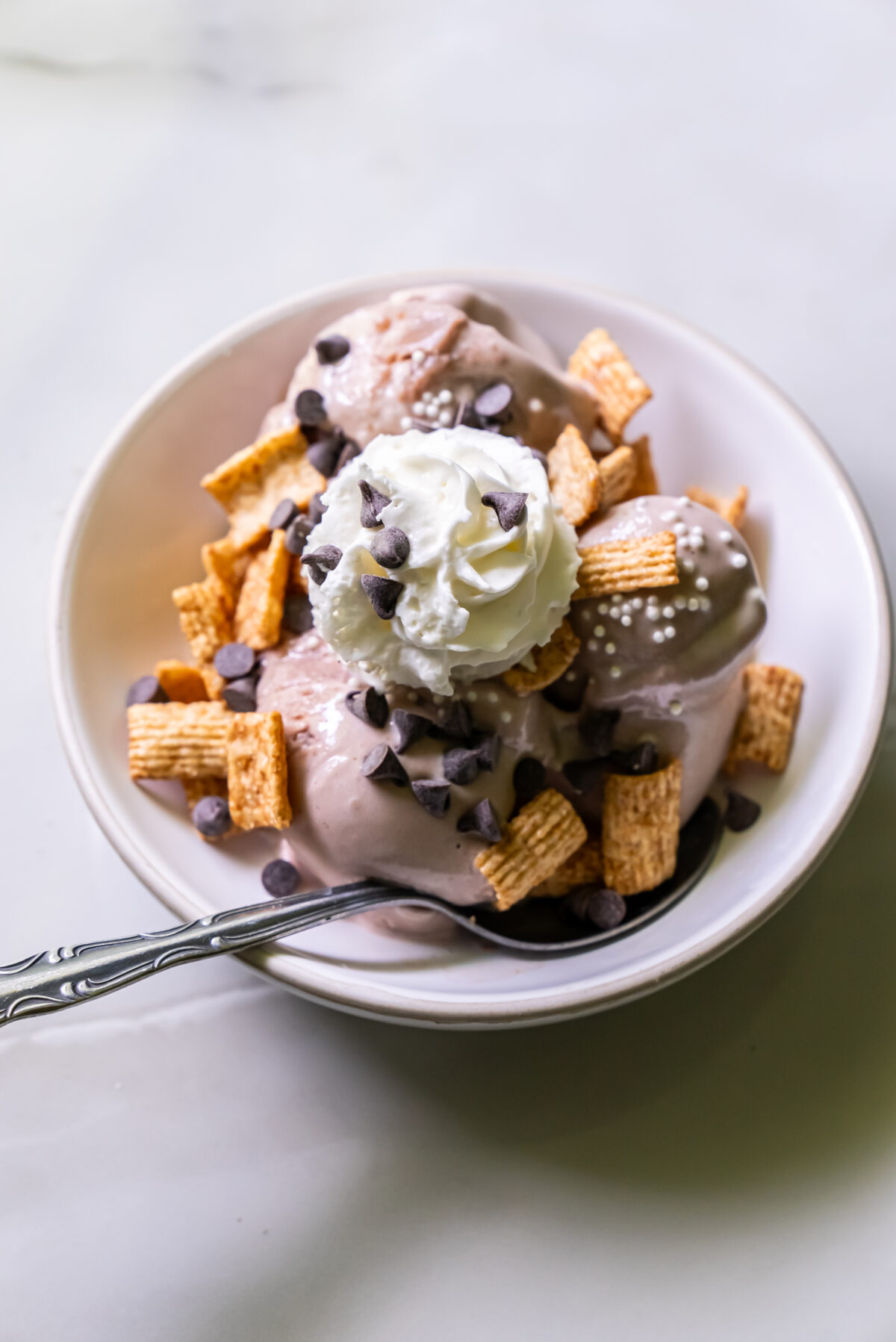 A bowl of ice cream topped with whipped cream, chocolate chips, and cereal pieces, with a spoon resting on the bowl's edge.