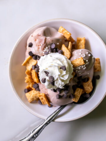 A bowl of Ninja Creami chocolate protein frozen yogurt topped with cereal pieces, chocolate chips, whipped cream, and white sprinkles on a marble surface.