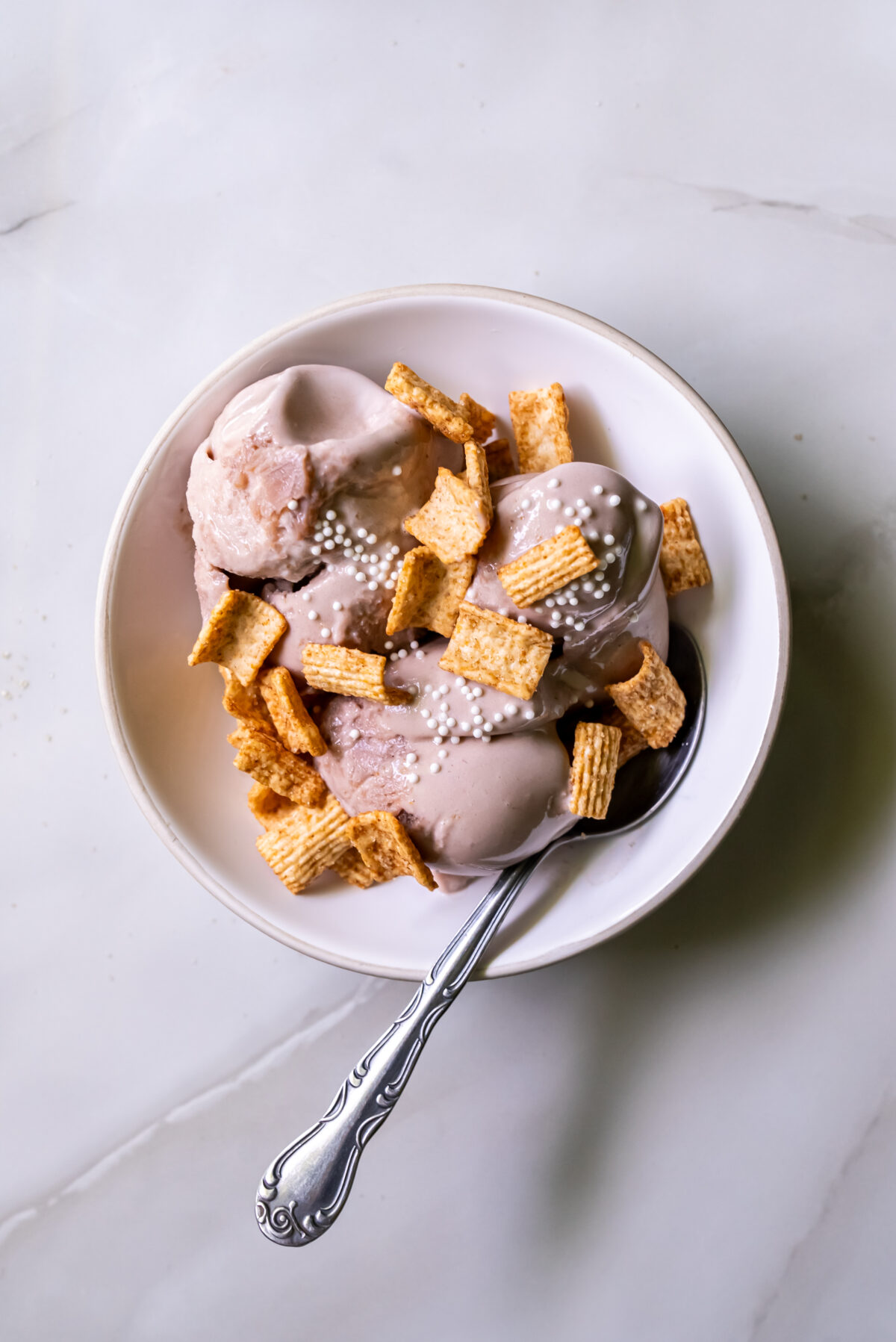 A bowl of ice cream topped with cereal pieces and small white sprinkles, with a spoon resting inside the bowl on a marble surface.