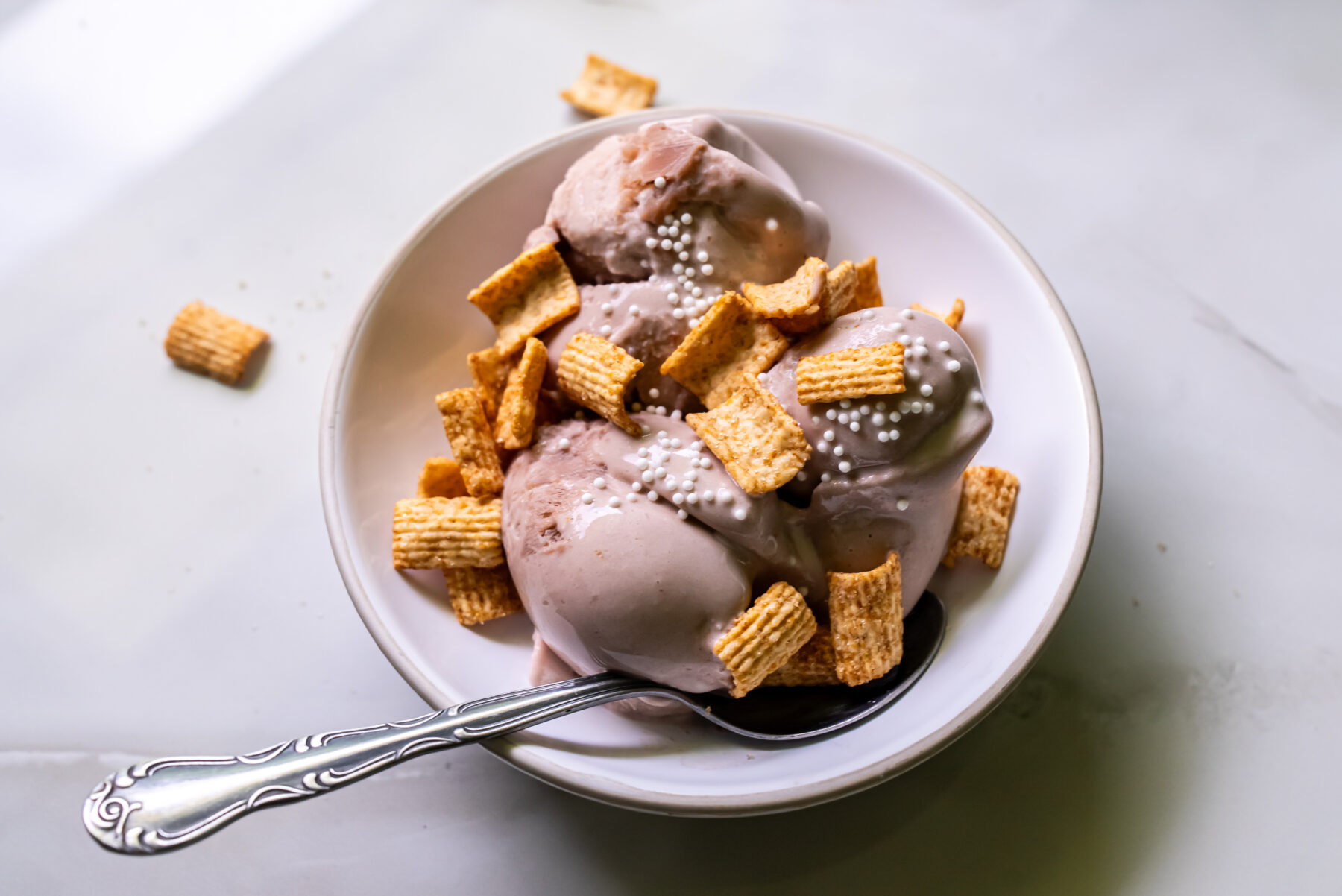 A bowl of ice cream topped with cereal pieces and small white sprinkles, with a spoon placed inside the bowl, on a light-colored surface.