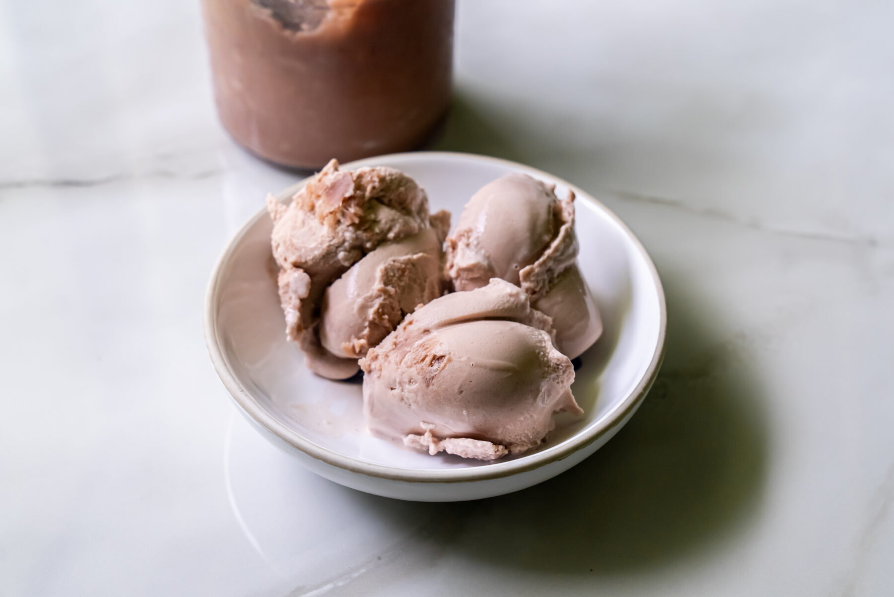 Three scoops of brown ice cream in a white bowl on a marble surface.