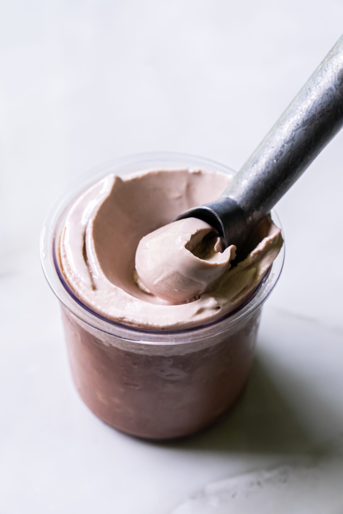 A scoop of chocolate ice cream in a clear plastic cup, with a metal ice cream scoop resting on top.