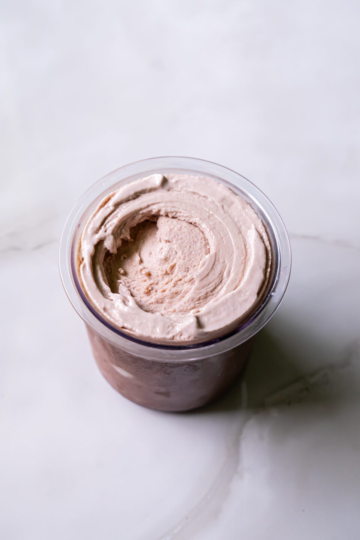 A plastic container filled with creamy, light brown ice cream, viewed from above, placed on a white surface.