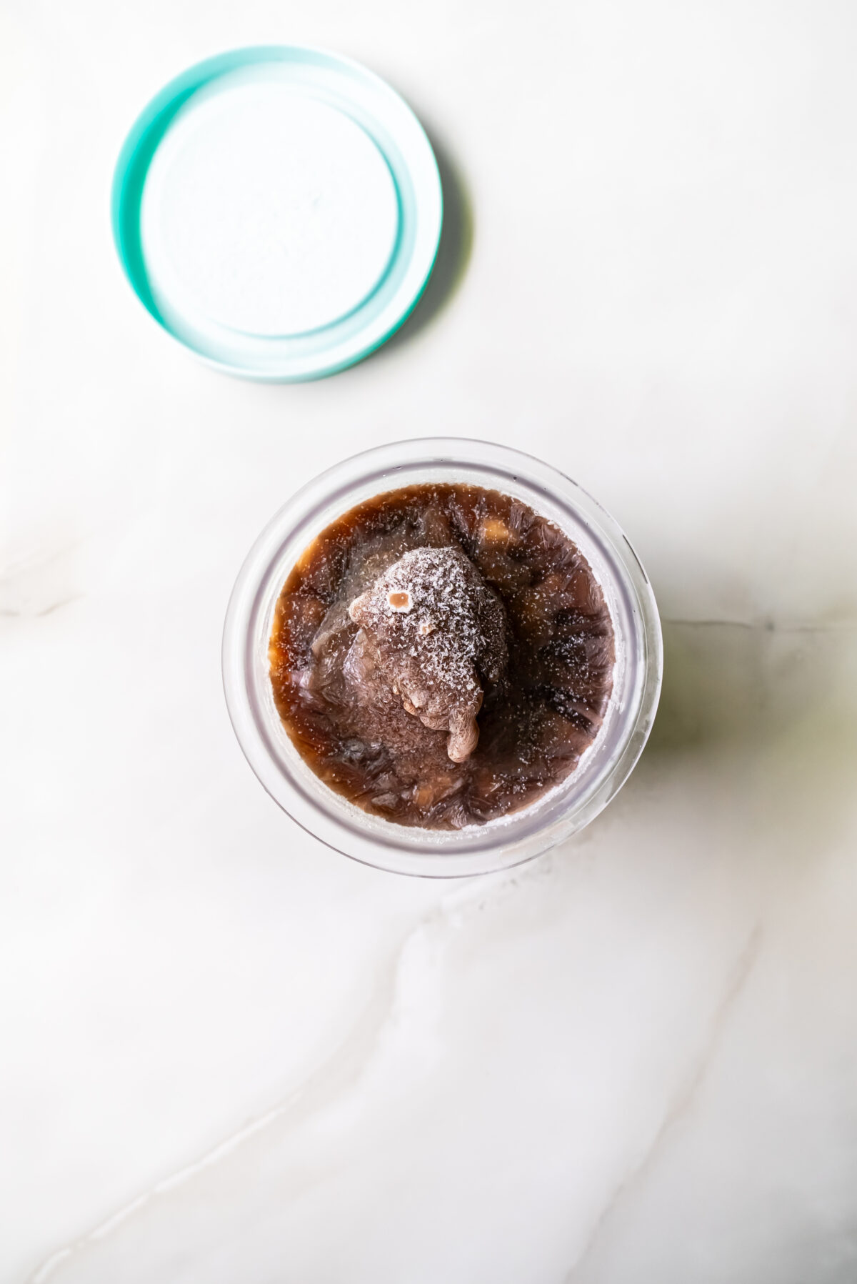 A glass container filled with a frozen brown beverage with the lid placed beside it on a white surface.