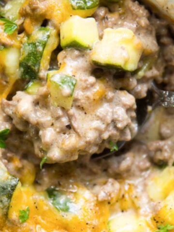A close-up view of a spoon scooping a cheesy ground beef and zucchini casserole, one of 26 filling low carb dinners, garnished with parsley.