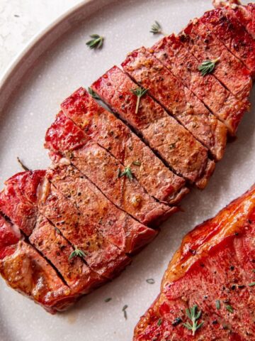 A close-up of a cooked, sliced steak garnished with sprigs of thyme, served on a gray plate, inviting you to savor the succulent flavors in the classic debate of Sirloin vs New York Strip.