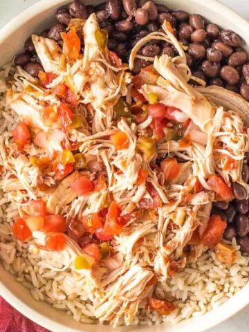 A bowl filled with shredded chicken, black beans, rice, and salsa is surrounded by a red napkin, a fork, cilantro, a lime half, and a small dish of sour cream on a light-colored surface. Ideal for one of your 20 dinners this month.