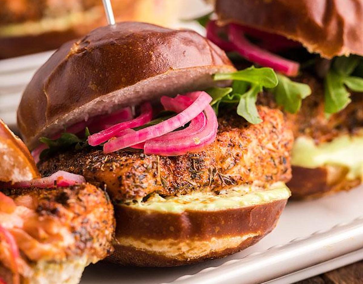 A close-up of a sandwich with a well-seasoned meat patty, pickled red onions, greens, and sauce, all inside a pretzel bun sits on a white plate. This could easily stand out among 25 seafood dishes at any gathering.