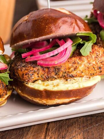 Three seasoned chicken sliders with arugula, pickled onions, and sauce sit on a white rectangular plate on a wooden table, standing out among 23 seafood dishes.