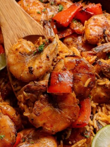 Close-up of a skillet containing spiced shrimp with red bell peppers, rice, and lime wedges, being stirred with a wooden spoon. The vibrant dish promises a burst of flavors, perfect for savoring in every bite.