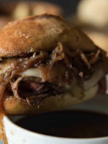 A close-up of a French dip sandwich with melted cheese and layers of caramelized onions, partially dipped in a cup of au jus.