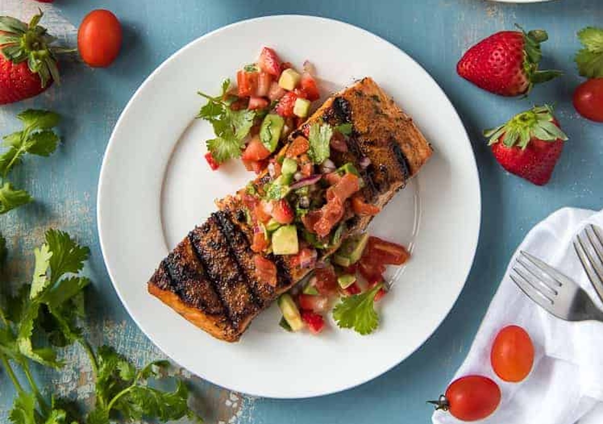 Grilled salmon topped with a mix of diced vegetables and herbs on a white plate, garnished with whole strawberries and cherry tomatoes. A perfect addition to your favorite recipes, forks are placed near the plate.