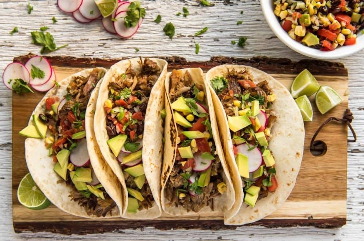 Four tacos filled with shredded meat, diced avocados, radish slices, chopped cilantro, and other toppings are arranged on a wooden board. A bowl of corn salsa and lime wedges are beside them. Perfect for a quick meal inspired by 11 instant pot recipes.