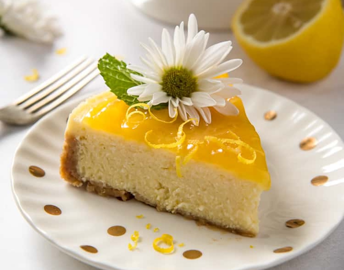A slice of cheesecake with a vibrant yellow topping, garnished with a white daisy and mint leaves, served on a plate adorned with delicate gold dots. In the background, a lemon and fork await your indulgence—a perfect 10 in dessert presentation.
