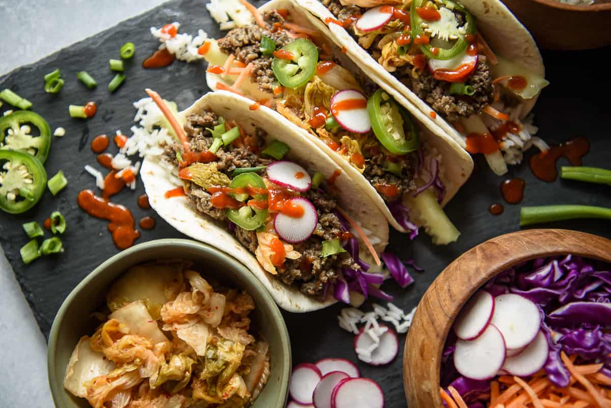 Three tacos filled with seasoned beef from our 20 beef recipes collection, sliced radishes, jalapeños, and drizzled with sauce, served on a slate board with a side dish of kimchi and a bowl of sliced radishes and purple cabbage.
