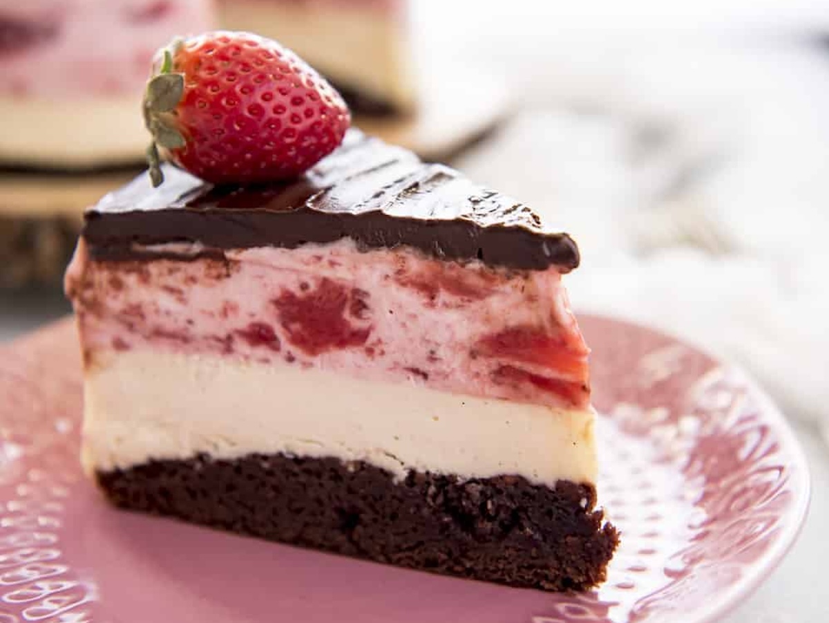 A slice of layered dessert on a pink plate. It has a chocolate base, white middle layer, strawberry layer, and is topped with chocolate and a whole strawberry—perfect for a 10-minute indulgence.