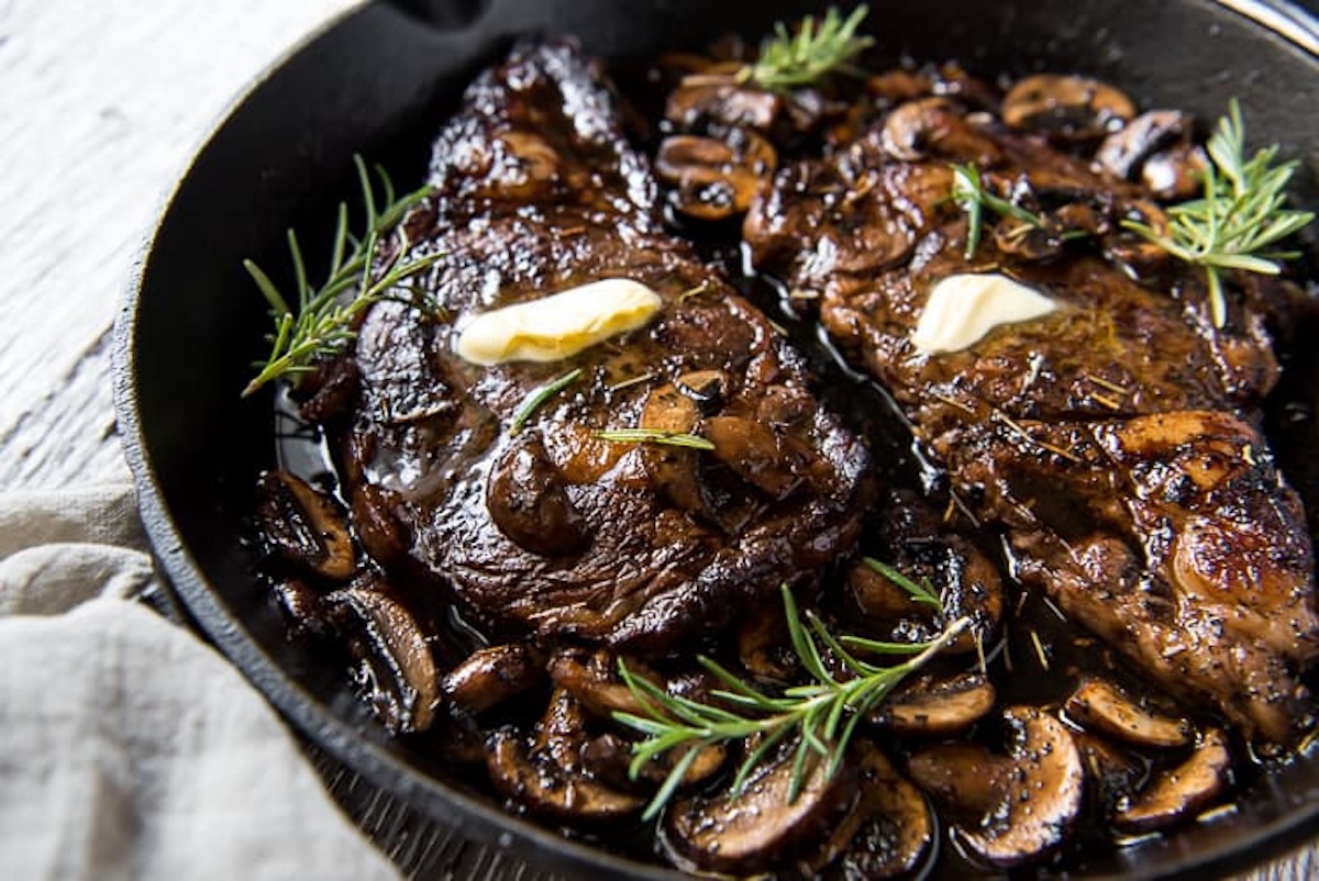 A skillet with two cooked steaks topped with butter, surrounded by sautéed mushrooms and garnished with sprigs of rosemary from one of our top 20 steak recipes.