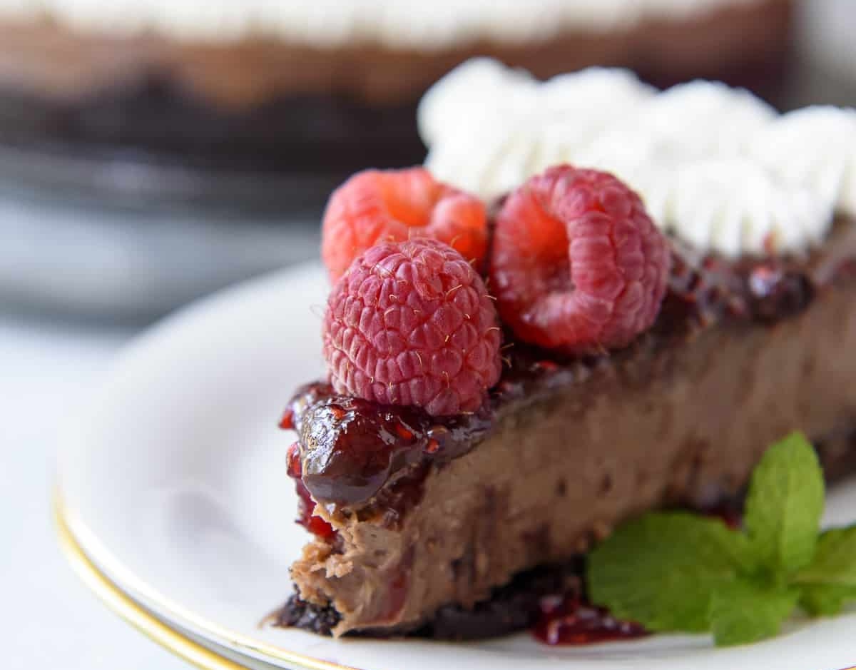 A decadent slice of chocolate cheesecake topped with fresh raspberries, luscious whipped cream, and sprigs of mint leaves on a pristine white plate awaits.