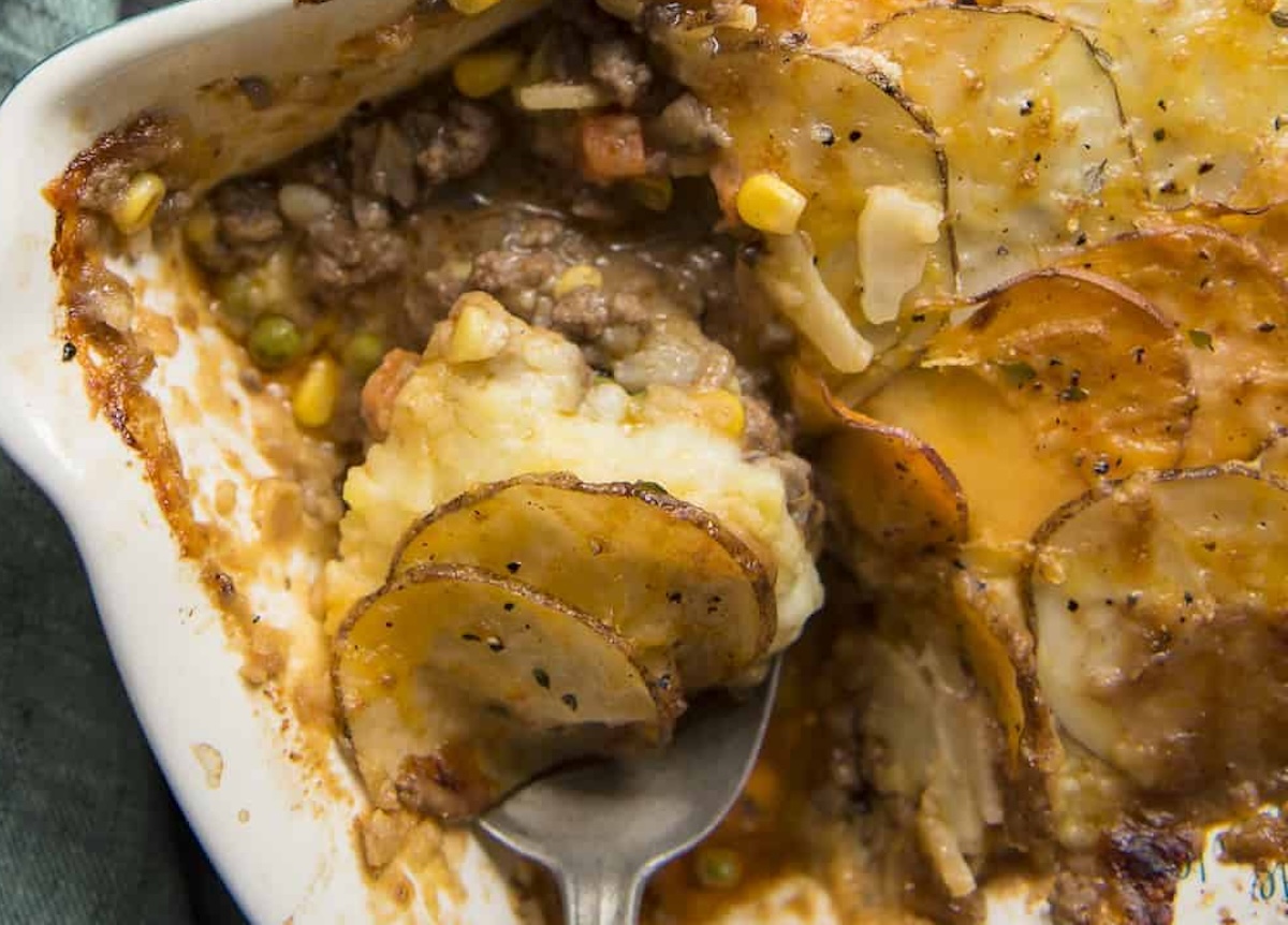 Close-up of a baked casserole dish with a serving spoon. This savory delight, one of the 13 potato dishes to try, contains a layered mix of sliced potatoes, ground meat, corn, and peas.