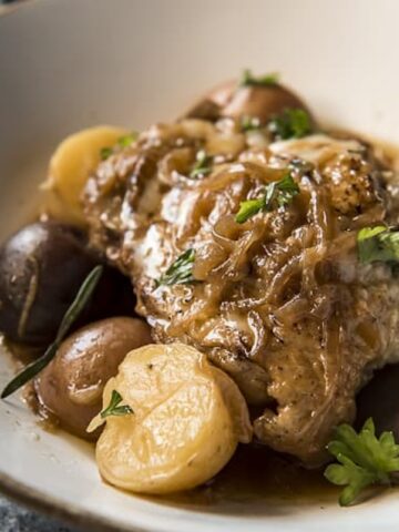 A dish of braised chicken, enhanced with 12 onion pieces, served with various small potatoes and garnished with herbs in a white bowl.