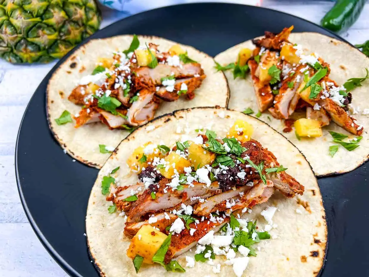 Three tacos with grilled chicken, pineapple chunks, cilantro, and cheese on a black plate – a delightful addition to your collection of go-to recipes.
