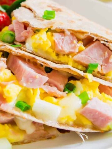 A plate with two quesadillas filled with ham, scrambled eggs, cheese, and green onions. Sliced strawberries are visible in the background, making it one of the 20 breakfasts you’ll want to try.