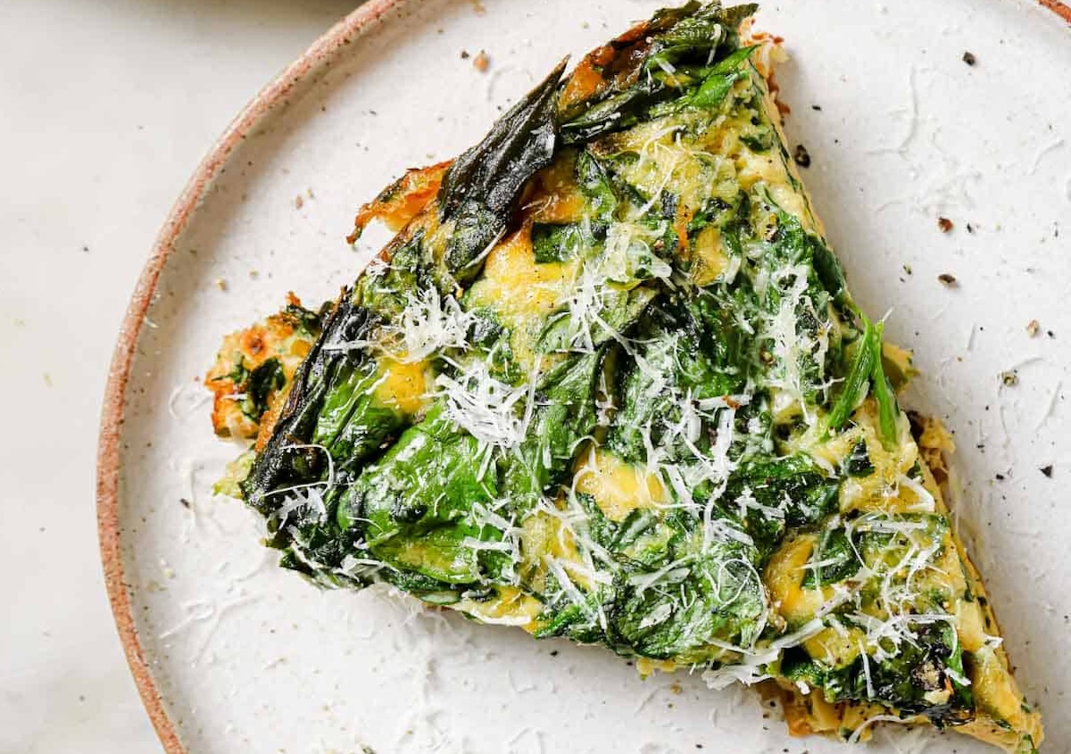 A triangular slice of veggie spinach frittata garnished with grated cheese, served on a white plate.
