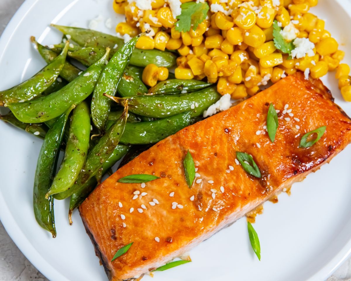 A plate featuring a grilled salmon fillet garnished with sesame seeds and green onions, served with a side of sautéed sugar snap peas and corn topped with cheese and herbs—perfect for when you're considering 10 air fryer fixes to elevate your meals.