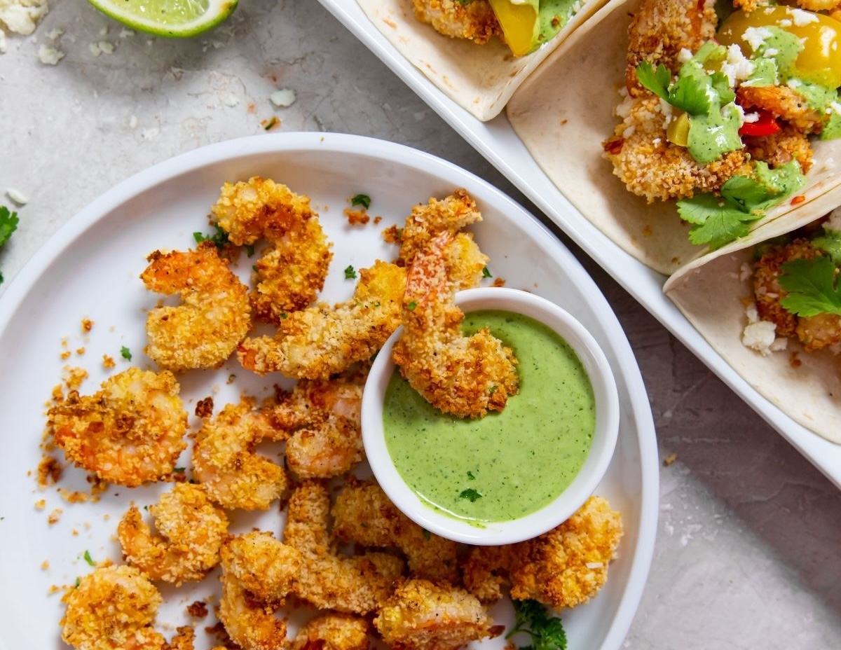 A plate of breaded shrimp next to a small bowl of green dipping sauce, with tacos filled with shrimp and garnished with cilantro in the upper corner. Lime slices are visible in the background—just one example from our collection of 10 air fryer recipes for delicious and healthier meals.