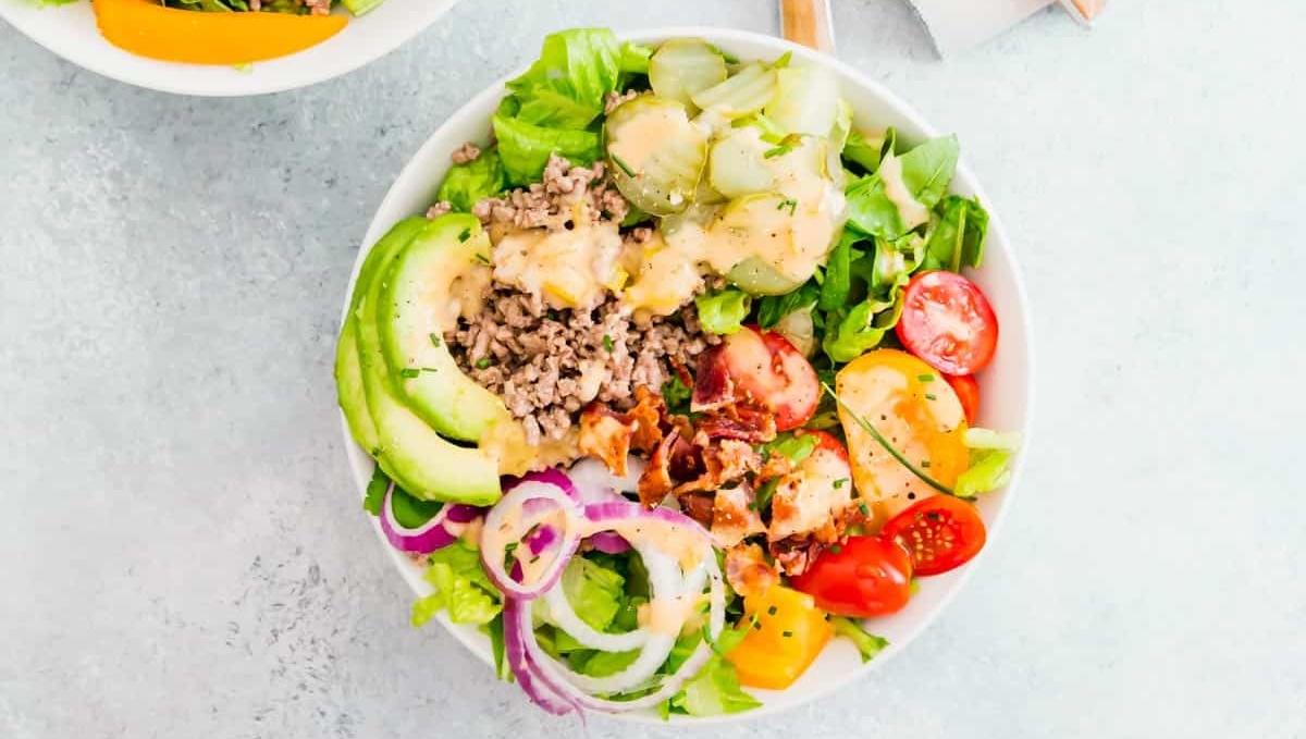 A bowl of salad featuring lettuce, ground meat, avocado slices, cucumber slices, cherry tomatoes, red onion rings, and a creamy dressing on top—one of the 10 must-try recipes for a fresh and wholesome meal.