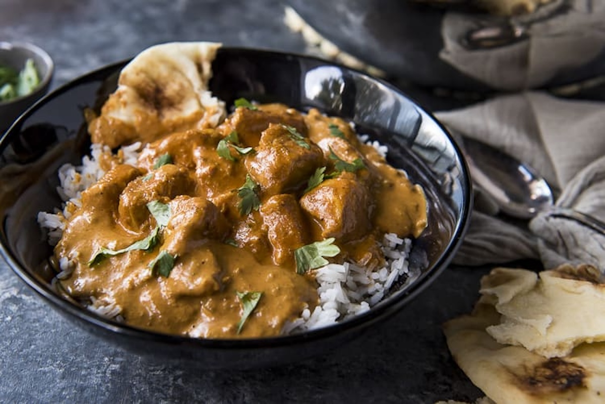 A black bowl filled with rice topped with chicken curry, garnished with cilantro, and accompanied by naan bread on a dark textured surface—one of the most delightful meals from our 19 dinners collection.