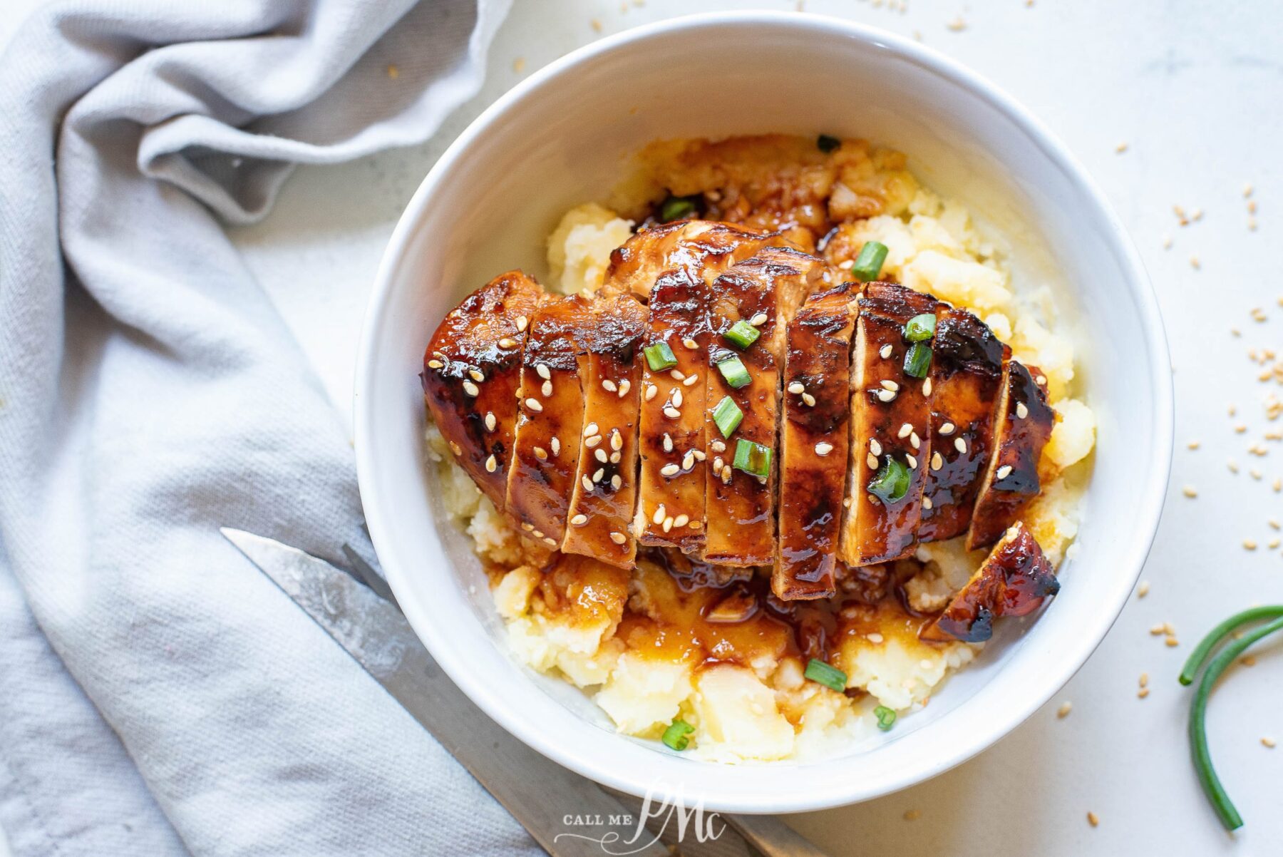 A white bowl containing 13 slices of teriyaki chicken breast garnished with sesame seeds and green onions, served over mashed potatoes. A knife and a cloth napkin are placed beside the bowl.