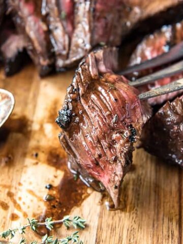 Close-up of a piece of cooked steak being held by a fork on a wooden cutting board, with a spoon and herbs nearby. Perfect for one of your 14 low carb dinners, the steak appears juicy and seasoned, displaying a medium-rare doneness.