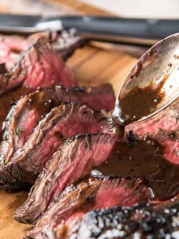 Slices of medium-rare beef steak being drizzled with a dark sauce, displayed on a wooden cutting board with a knife in the background.