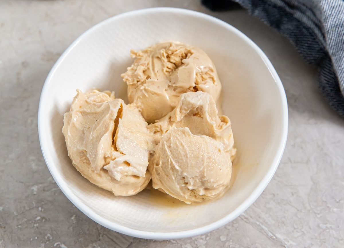 A white bowl containing three scoops of light brown Ninja Creami ice cream on a textured surface, with a dark cloth partially visible in the background.