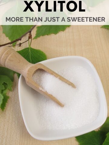 A small dish filled with granulated xylitol, accompanied by a wooden scoop, is placed on a wooden surface with green leaves around. The text above the image reads, "Xylitol: More than just a sweetener.