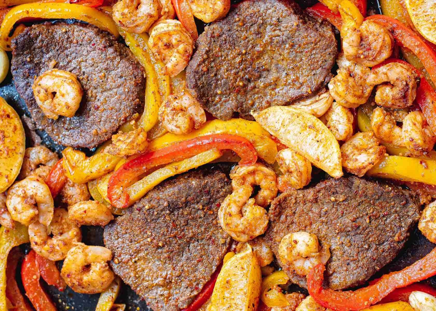 A close-up of a cooked dish featuring seasoned beef slices, shrimp, red and yellow bell peppers, and potato wedges, perfect for adding to your collection of 12 sheet pan dinners.