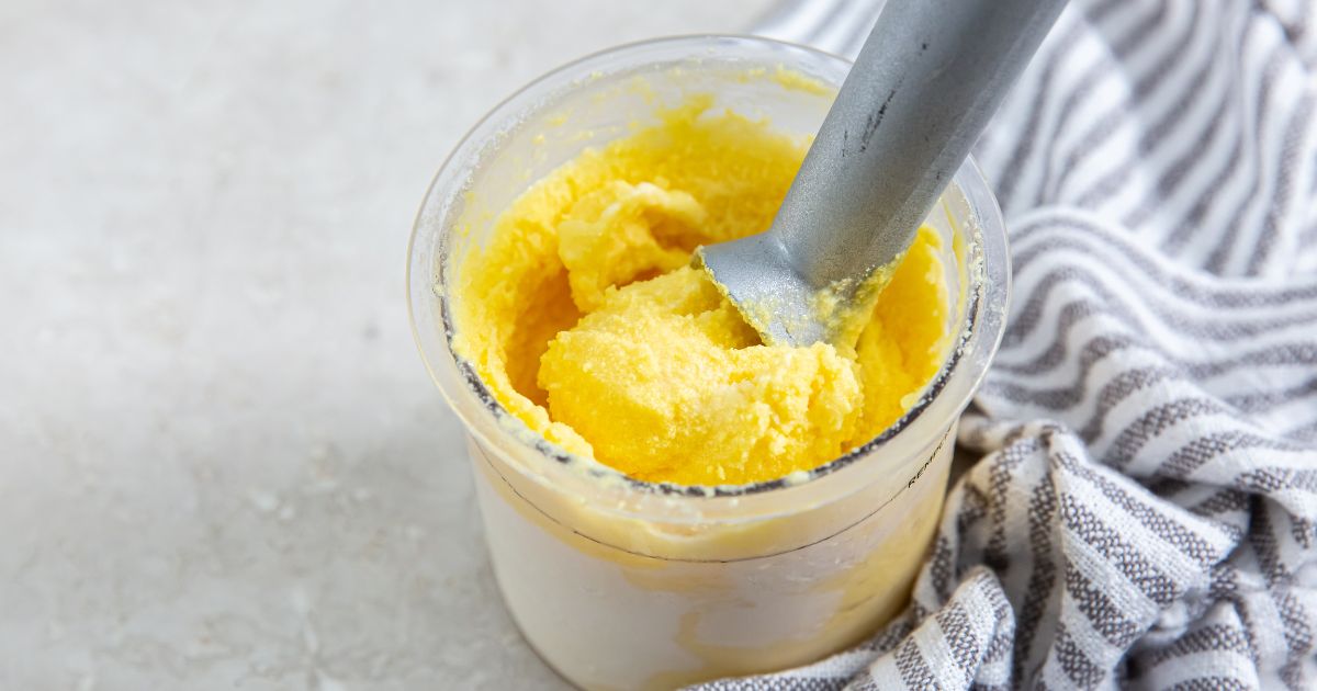 A glass jar of freshly churned yellow butter with a metal spatula inside, placed next to a striped cloth.