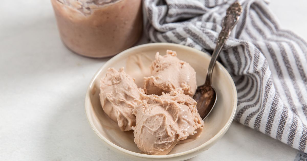 A bowl of light brown ice cream with a metal spoon, next to a jar and a striped cloth on a white surface.