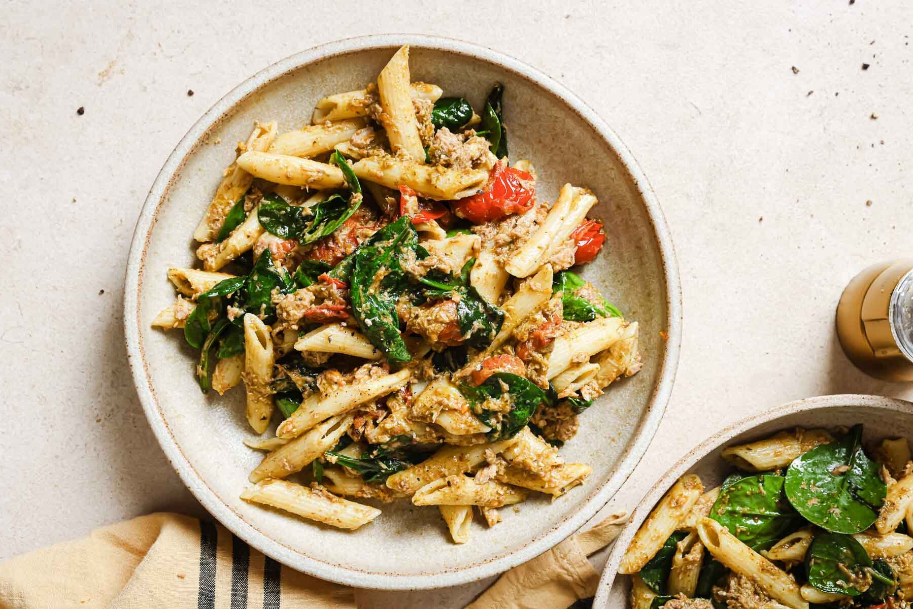 A bowl of penne pasta with spinach, cherry tomatoes, and pesto sauce on a light-colored surface.