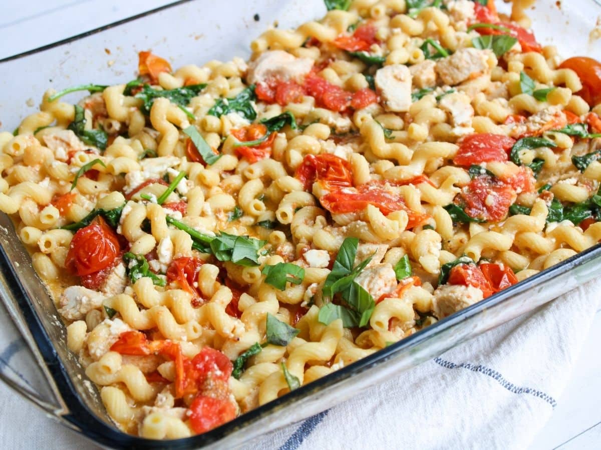 A glass baking dish filled with cooked pasta, cherry tomatoes, spinach, and crumbled tofu or cheese, topped with fresh basil.