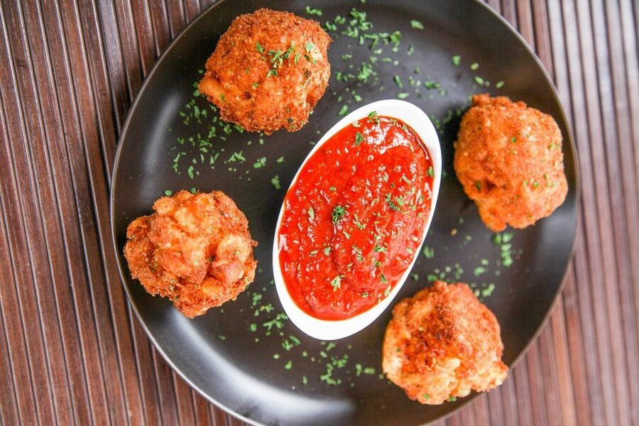 A black plate with four fried arancini balls surrounding a dish of red marinara sauce, garnished with chopped herbs.