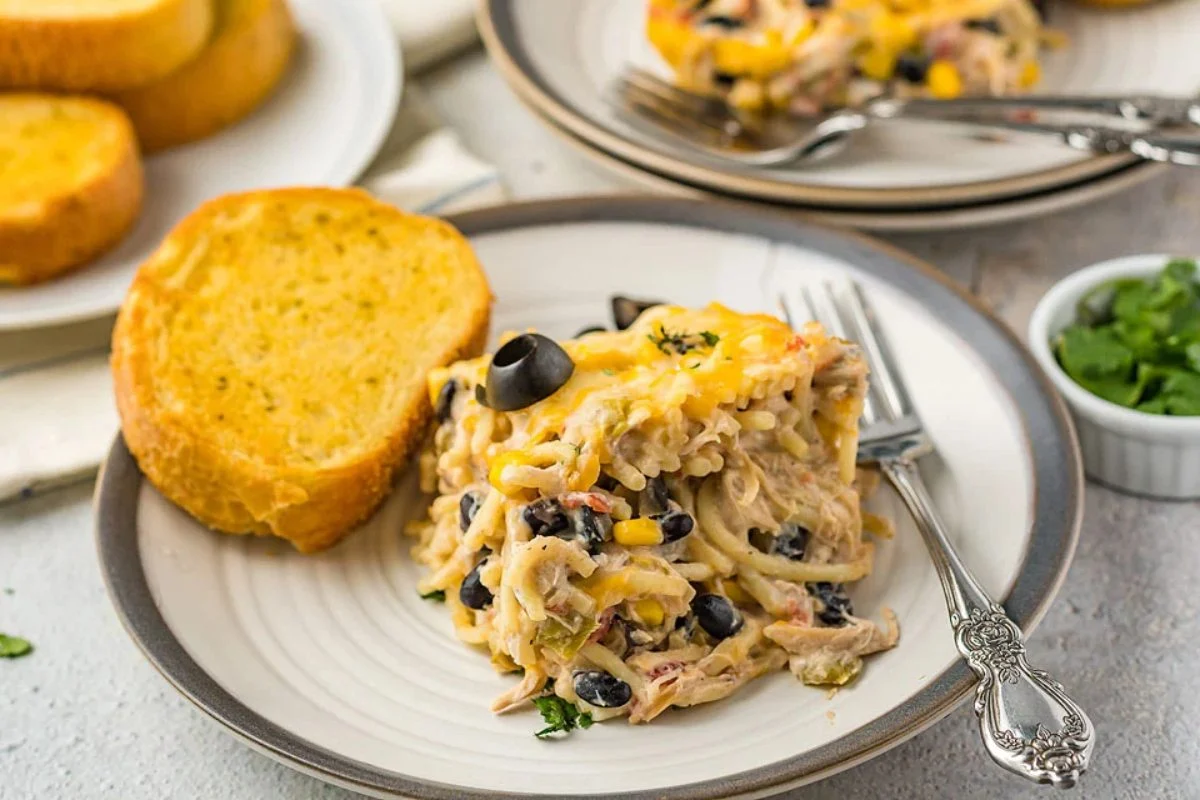A plate of cheesy pasta bake with black olives and corn, served with a slice of garlic bread. Another plate and a bowl of cilantro are visible in the background, reminiscent of one of those hearty 15 chicken dinners that bring comfort to the table.