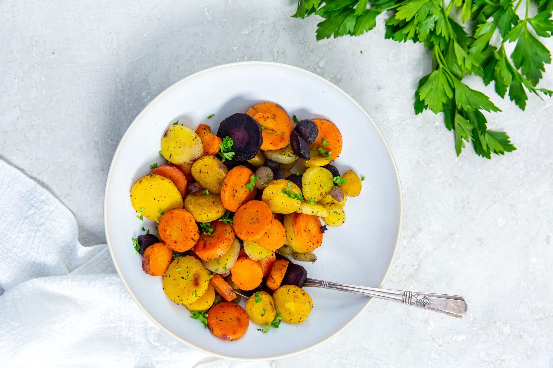 A white bowl filled with sliced and roasted colorful carrots, garnished with herbs. A silver fork rests inside the bowl. A bunch of parsley is placed nearby on a light-colored surface.