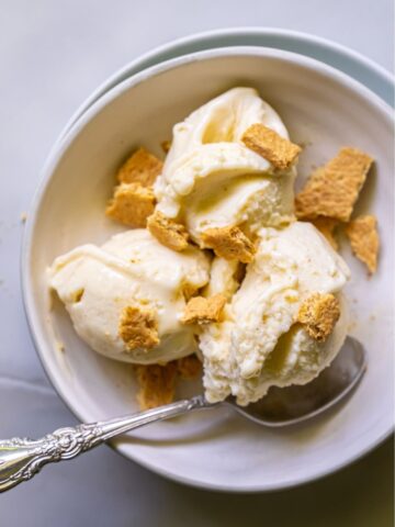 A bowl of banana cheesecake ice cream topped with crumbled graham crackers, with an ornate spoon inside the bowl, placed on a light-colored surface.